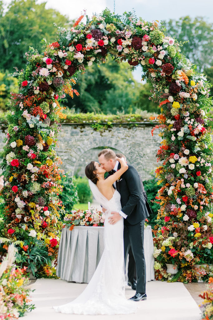 Bride and Groom kiss in front flower arch at their outdoor wedding ceremony in The Walled Garden at Adare Manor